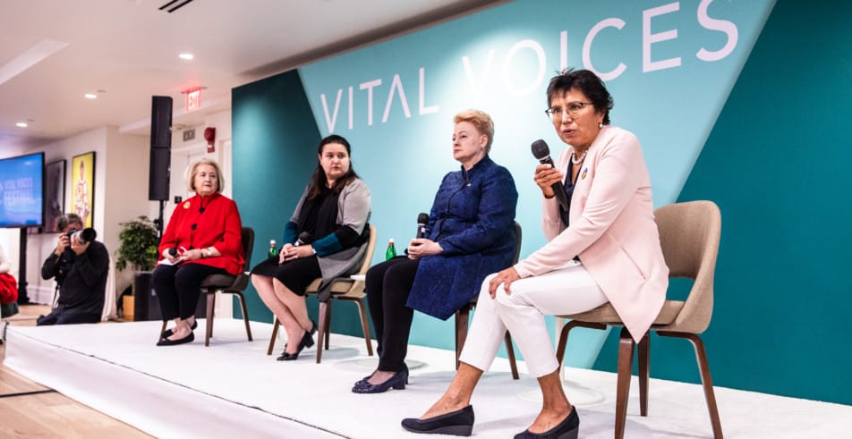 Four women sit on a stage in front of the Vital Voices Logo
